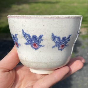 Puede incluir: Un cuenco de cerámica blanca con diseños florales azules y centros rojos. El borde tiene un tinte rosado. El cuenco se sostiene en una mano, mostrando su forma. El diseño se repite alrededor del cuenco.