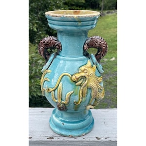 May include: A turquoise ceramic vase featuring a relief-molded yellow dragon design wrapping around the body. The vessel has two dark brown handles shaped like stylized creatures and a wide, flared rim with a visible glaze finish.