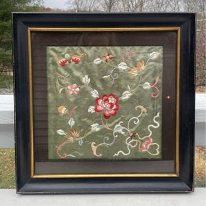 May include: A framed embroidered artwork featuring a floral design with pink, red, and white flowers on a green background. The frame is black with a gold inner border.