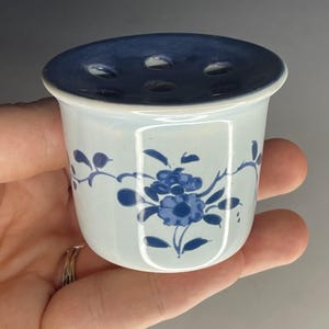 May include: A small, white ceramic flower frog with a dark blue glazed top featuring five circular holes. The exterior of the vessel is decorated with a hand-painted blue floral pattern consisting of blossoms, leaves, and vines.