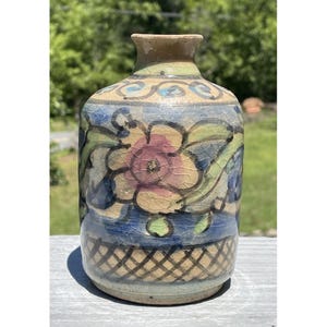 May include: A ceramic vase with a floral design. The vase features a pink and yellow flower with green leaves, set against a blue and beige background. The base has a crosshatch pattern. The vase is approximately 7 inches tall.