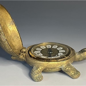 Könnte beinhalten: Eine goldfarbene, vintage-förmige Schildkrötenuhr mit einem aufklappbaren Panzer. Das Zifferblatt hat römische Ziffern und eine transparente Abdeckung. Die Schildkröte hat grüne Akzente am Kopf.