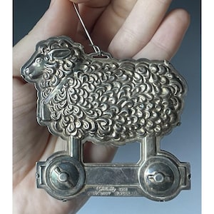 Może przedstawiać: Srebrna ozdoba w kształcie owcy z szczegółowym, teksturowanym wzorem. Ozdoba ma pętlę do zawieszenia i jest zamontowana na podstawie z kółkami. Widoczny jest tekst "1981, The Kirkhoff Silverplate".