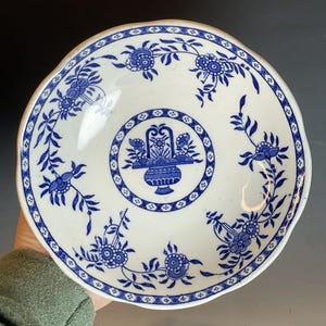 Könnte beinhalten: Eine weiße Keramikschüssel mit einem blauen und weißen Blumendesign. Die Schüssel hat einen gewellten Rand und ein zentrales Design mit einem Blumenkorb.