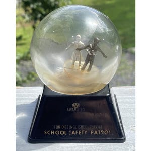 May include: A vintage snow globe with two miniature figures inside, set on a black base. The globe features a clear glass sphere with a yellowed liquid and a base that reads "FOR DISTINGUISHED SERVICE SCHOOL SAFETY PATROL."