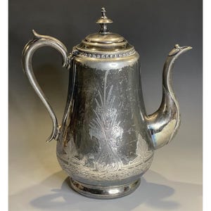 May include: Silver-plated coffee pot with a curved handle and spout. The pot features ornate floral and leaf engravings on the body and a decorative lid with a small finial. The pot sits on a round base.