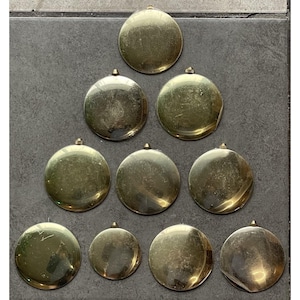 May include: A collection of ten tarnished, round, brass-colored pendants with small loops at the top. The pendants vary slightly in size and are arranged on a dark gray surface. The pendants have a vintage or antique appearance.