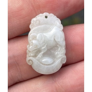 May include: White jade pendant carved with a detailed depiction of a dog. The pendant is oval shaped and has a loop at the top for hanging.