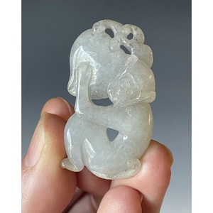May include: A carved jade pendant in a light green hue. The pendant features a detailed design of a figure holding a fruit or object, with intricate carvings on the head and body. The jade has a smooth, polished finish.