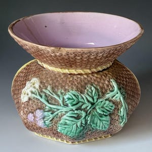 May include: A decorative ceramic planter with a woven basket design. The planter features a light purple interior and is adorned with green leaves, white flowers, and purple berries. The exterior is brown with yellow accents.
