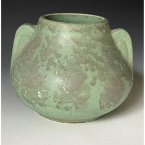May include: A light green ceramic vase with two side handles and a mottled, textured surface pattern in a darker shade of green. The vessel features a wide, rounded body and a circular opening at the top, showcasing a matte finish throughout.