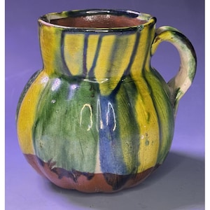 Könnte beinhalten: Eine handbemalte Keramikkanne mit gelber, grüner und blauer Glasur. Die Kanne hat einen runden Körper und einen Henkel.