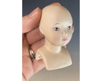 Op de afbeelding: Een porseleinen poppenhoofd met geschilderde zwarte ogen, roze lippen en een kaal hoofd. Het poppenhoofd is bevestigd aan een witte basis met de woorden "Made in Japan" erop gedrukt.