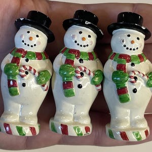 以下が含まれることがあります： 3体の陶器製の雪だるまの置物。各雪だるまは黒いシルクハット、緑と赤のマフラー、緑のミトンを着用しています。彼らはキャンディケインを持ち、ニンジン鼻と石炭の目で描かれた顔をしています。