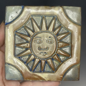 May include: Square ceramic tile with a sun face design. The tile features a raised sun with a face in the center, surrounded by pointed rays. The color palette includes beige, brown, and blue accents. The tile is approximately 10 cm square.