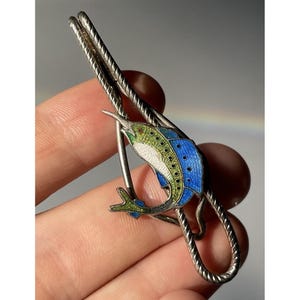May include: A silver-toned metal tie clip featuring a rope-textured bar and an enameled marlin fish motif. The fish is decorated with vibrant blue, green, and white enamel detailing, showcasing a pointed bill and a curved body shape.