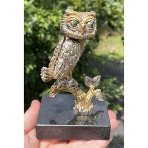 May include: A detailed silver and gold-toned owl figurine with green eyes, perched on a branch. The owl is mounted on a dark gray square base. The sculpture is held in a person's hand, with a blurred green background.