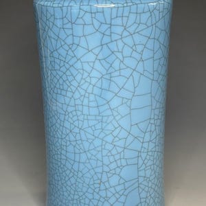 May include: A cylindrical ceramic vase with a light blue glaze and a prominent dark grey crackle pattern covering the entire exterior surface. The vase features a slightly flared rim and a smooth, rounded base, showcasing a distinct network of fine lines.