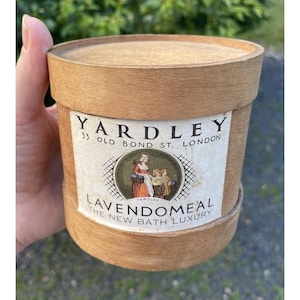 May include: A cylindrical light brown wooden container with a lid. The label reads YARDLEY 33 OLD BOND ST. LONDON LAVENDOMEAL THE NEW BATH LUXURY and features a circular illustration of two figures in period clothing in a garden setting.