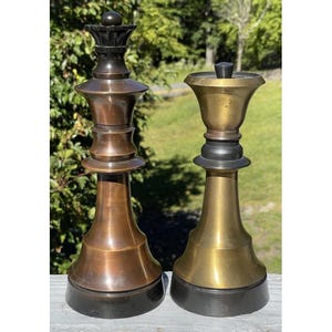 May include: Two large chess pieces standing side by side. The piece on the left is a dark bronze queen with a crown top. The piece on the right is a gold-toned queen with a smooth, flat top. Both pieces feature a wide, circular black base.