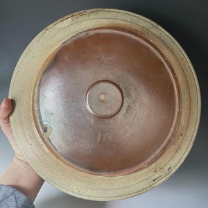 Może przedstawiać: Okrągła ceramiczna pokrywka z gładkim, ciemnobrązowym szkliwionym środkiem i wypukłym okrągłym uchwytem. Zewnętrzna krawędź wykonana jest z teksturowanej, jasnobeżowej kamionki z widocznymi niedoskonałościami i subtelnym pęknięciem na brzegu.