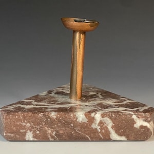 Puede incluir: Un cenicero de color cobre con una base triangular de mármol. El cenicero tiene un cuenco pequeño y un tallo largo. La base es de color marrón rojizo con vetas blancas. La base mide unos 10 cm de ancho.