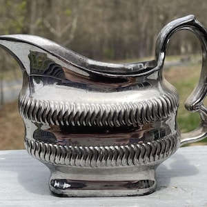 May include: A silver creamer with a rope-like design around the body and a handle. The creamer is made of metal and has a shiny finish.