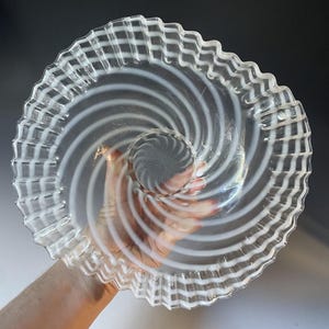 May include: Clear glass serving plate with a spiral design. The plate features a central, circular indentation and a wavy, textured rim. The design includes white, swirling lines radiating from the center.