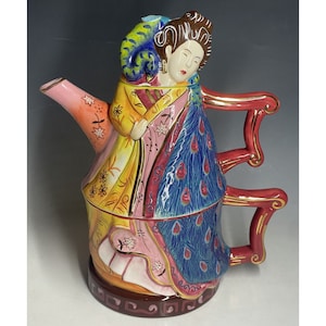 May include: A colorful ceramic teapot shaped like a woman in a traditional Asian dress. The teapot has a pink spout, a red handle, and a blue and green peacock design. The woman is wearing a yellow and pink robe with a gold trim.