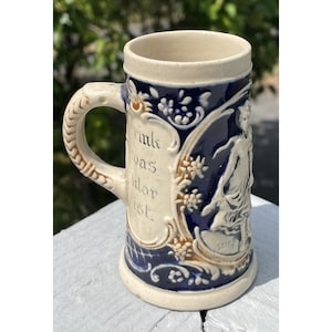 May include: A white ceramic beer stein with a blue and gold design. The stein features a raised design of a woman in a dress and flowers. The text "Trink was klar ist" is written on the stein.