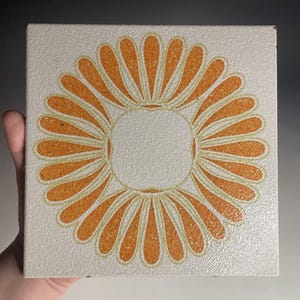 Puede incluir: Azulejo cerámico cuadrado con diseño floral. El azulejo presenta un círculo central rodeado de formas anaranjadas en forma de pétalo sobre un fondo blanco roto texturizado. El azulejo mide aproximadamente 15 cm cuadrados.
