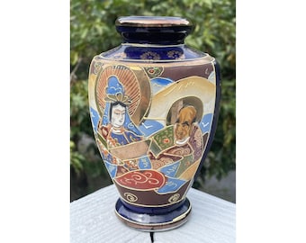 May include: A small, blue and gold ceramic vase with a Japanese-inspired design. The vase features a scene with two figures, one wearing a blue and gold robe and the other wearing a green and gold robe. The vase is decorated with intricate details and has a glossy finish.