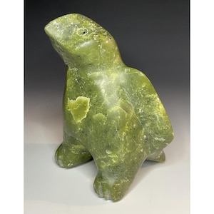 May include: A green stone sculpture of a seal. The seal is in a seated position with its head tilted upwards. The sculpture has a smooth, polished surface with natural variations in colour and texture. The sculpture is approximately 20 cm tall.