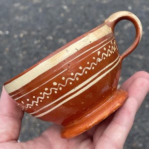 Może przedstawiać: Mały, ręcznie malowany ceramiczny kubek z uchwytem. Kubek jest głównie czerwonobrązowy z kremowymi paskami i białymi wzorami w kropki i faliste linie. Podstawa i uchwyt są również pomalowane na pomarańczowo i kremowo.