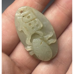 May include: A pale green jade pendant carved with a detailed design of a mythical creature. The pendant is oval shaped and has a smooth, polished surface.