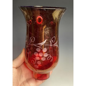 May include: A ruby red glass vase with a flared top and a round base. The vase features etched grape and vine designs. The glass has a glossy finish. The vase is held by a hand.
