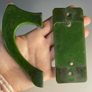 May include: Two dark green marbled objects, one a curved handle and the other a rectangular plate with two circular holes and two small metal studs. The pieces feature a smooth, polished finish with swirling patterns throughout the green material.