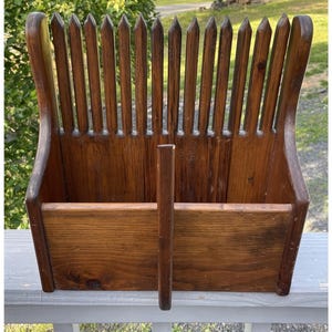 May include: A vintage wooden berry picker with a dark brown finish. The basket has a handle and a series of vertical tines at the top. The sides are curved, and the overall design is rustic and functional.