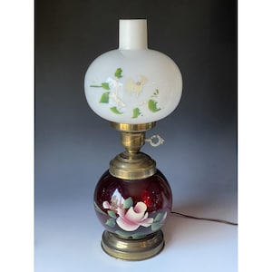 Peut inclure: Une lampe de table vintage avec un abat-jour globe blanc orné d'accents floraux verts et jaunes. La lampe présente une base en laiton et un corps en verre rouge foncé, également décoré d'un motif floral rose et blanc. Un cordon d'alimentation s'étend de la base.
