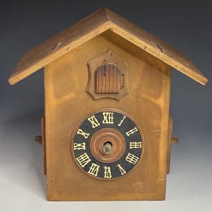 Op de afbeelding: Een houten koekoeksklok met een schuin dak en een ronde wijzerplaat. De wijzerplaat is zwart met gouden Romeinse cijfers. Boven de wijzerplaat bevindt zich een kleine, decoratieve deur. De klok is gemaakt van lichtbruin hout.
