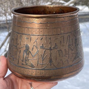 May include: A copper-colored, cylindrical vessel with a wide opening and a slightly tapered base. The exterior features etched figures and symbols, possibly depicting an ancient scene. The vessel is held in a hand.
