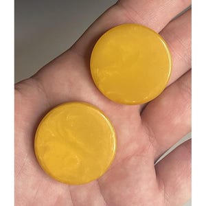 May include: Two round, golden-yellow objects with a marbled appearance. The objects are smooth and shiny, with a slight variation in color. The items are held in a hand, suggesting they are small and portable.