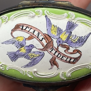 May include: A vintage oval-shaped compact with a green and white enamel design. Two purple and yellow birds are flying with a ribbon that reads "Amans Fidelle".