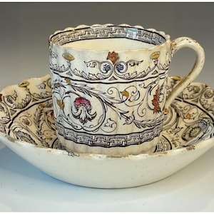 Może przedstawiać: Antyczny zestaw filiżanki i spodka. Kremowa filiżanka i spodek ceramiczny mają szczegółowy kwiatowy i geometryczny wzór w kolorze czarnym, czerwonym, żółtym i niebieskim. Filiżanka ma uchwyt i stoi na pasującym spodku z falistą krawędzią.