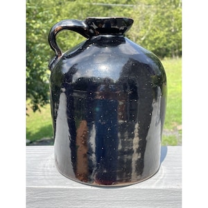 May include: A large, dark brown ceramic jug with a handle. The jug has a rounded body and a narrow neck. The handle is arched and attached to the top of the jug. The jug is glazed and has a glossy finish. The jug is sitting on a gray surface.
