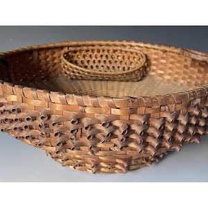 May include: A large, round, woven basket with a smaller, rectangular basket nested inside. The basket is made of light brown woven material with a textured, spiky design around the rim. The interior is a lighter shade of brown.