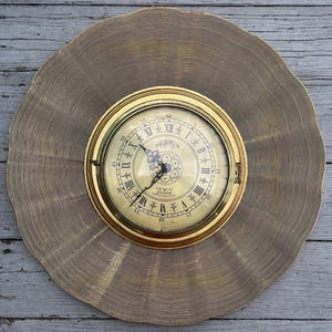 Puede incluir: Un reloj de estilo antiguo con marco dorado y esfera crema, con números romanos y agujas decorativas. El reloj está sobre un fondo texturizado, circular y dorado. El diámetro del reloj es de aproximadamente 25 cm.