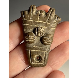 May include: A small, brown, carved pendant with a stylized design. The pendant features a rectangular base with a circular hole, two circular shapes, and a crown-like top. The pendant is approximately 2 inches long.