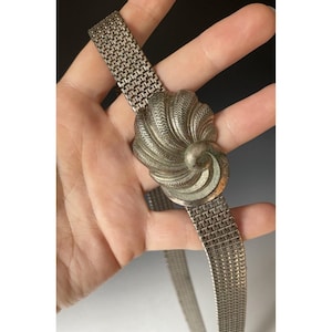 May include: A silver-toned metal belt with a decorative, swirling centerpiece. The belt features a mesh-like band and a detailed, textured design on the central element. The belt is held in a hand.