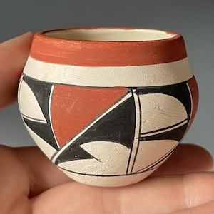 May include: A small, round, hand-painted ceramic pot with a red rim and geometric designs. The pot features a white base with black and red triangular and curved shapes. The interior is white, and the pot is held in a hand.
